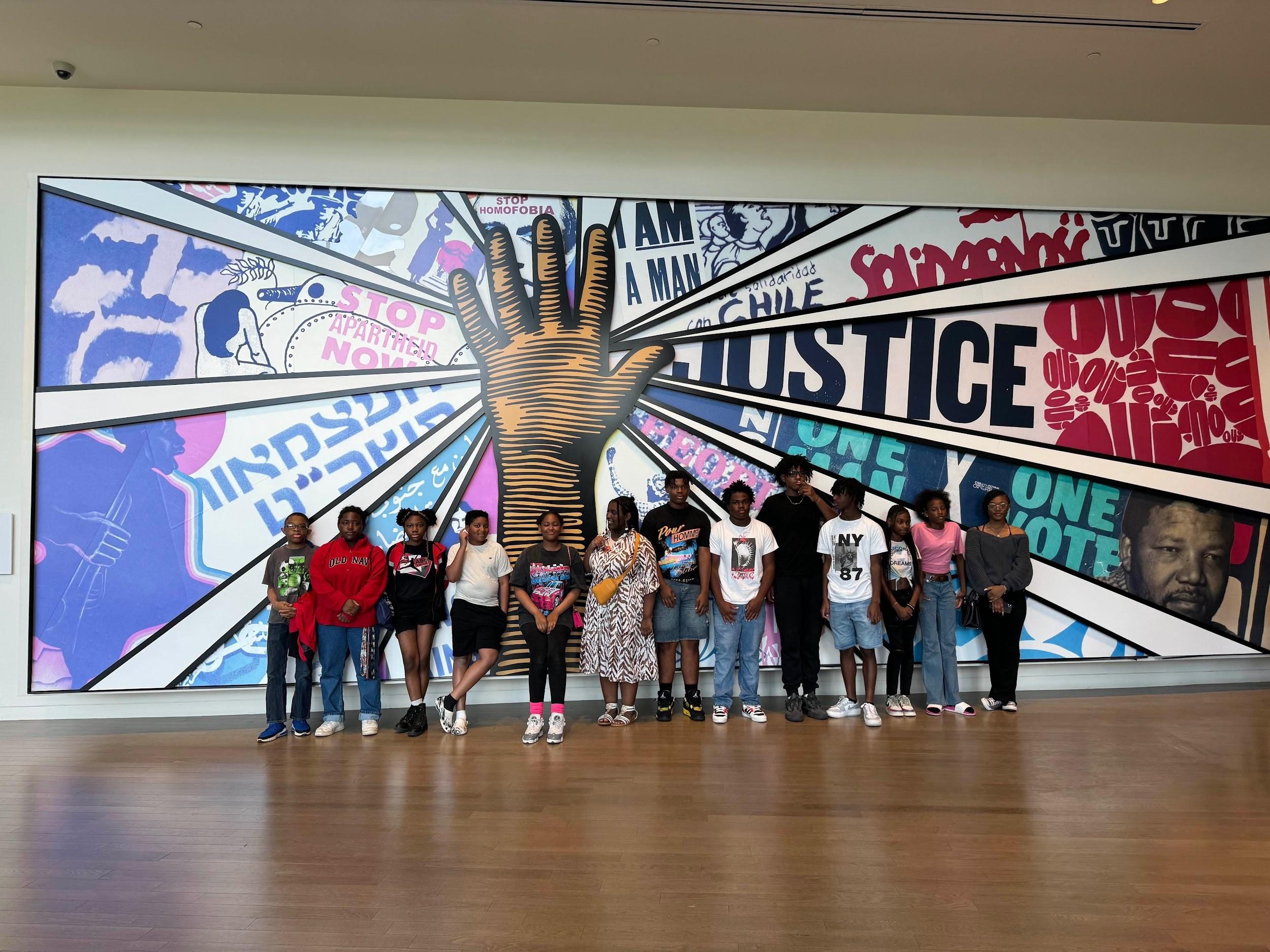 Group of children posing with raised hands in front of a colorful justice mural.