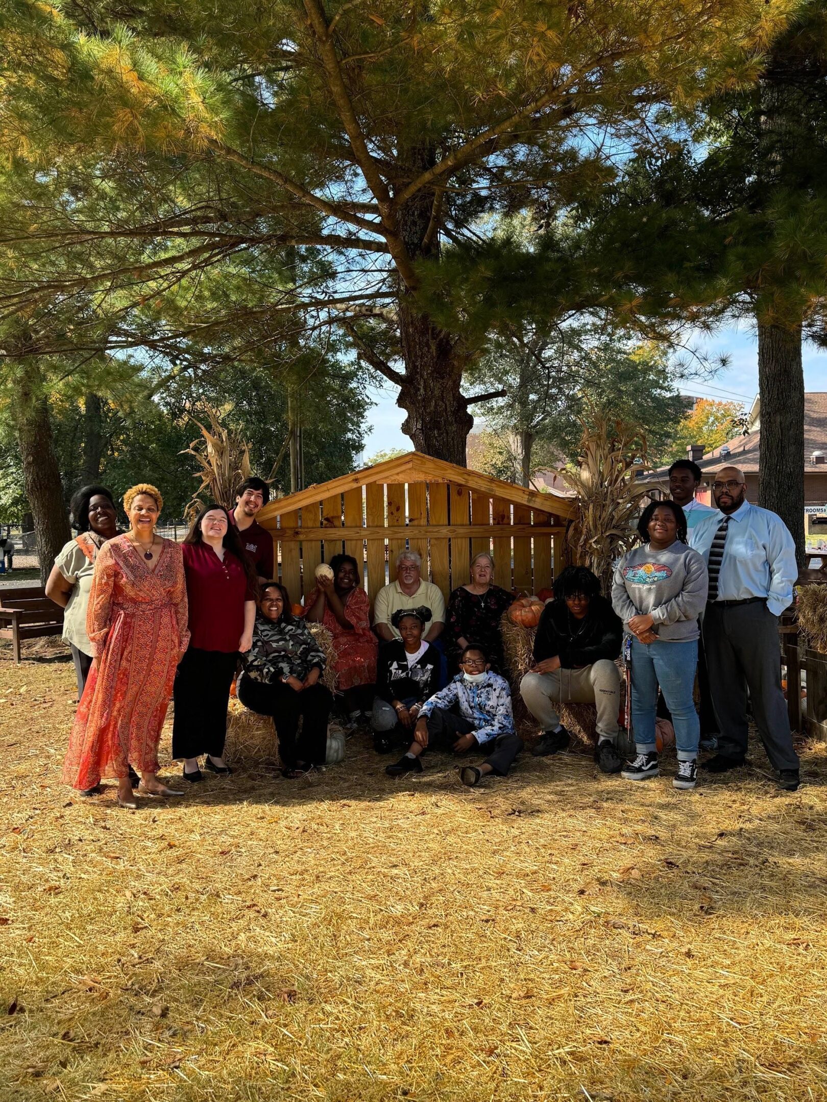 Group of people gathered outdoors around a decorated wooden structure under trees.