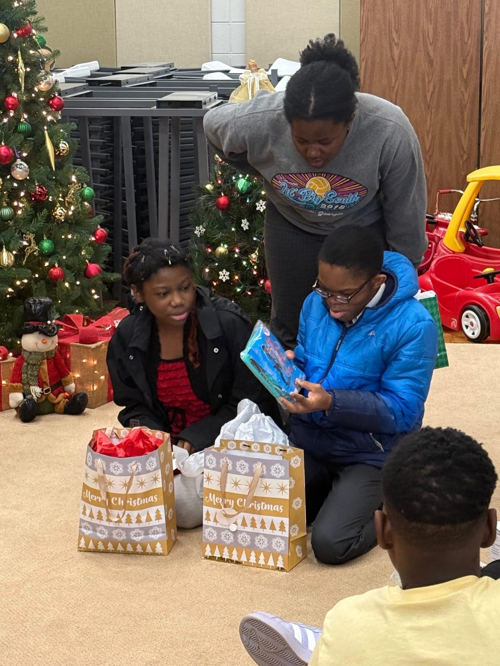 Children opening presents near a Christmas tree with an adult watching.