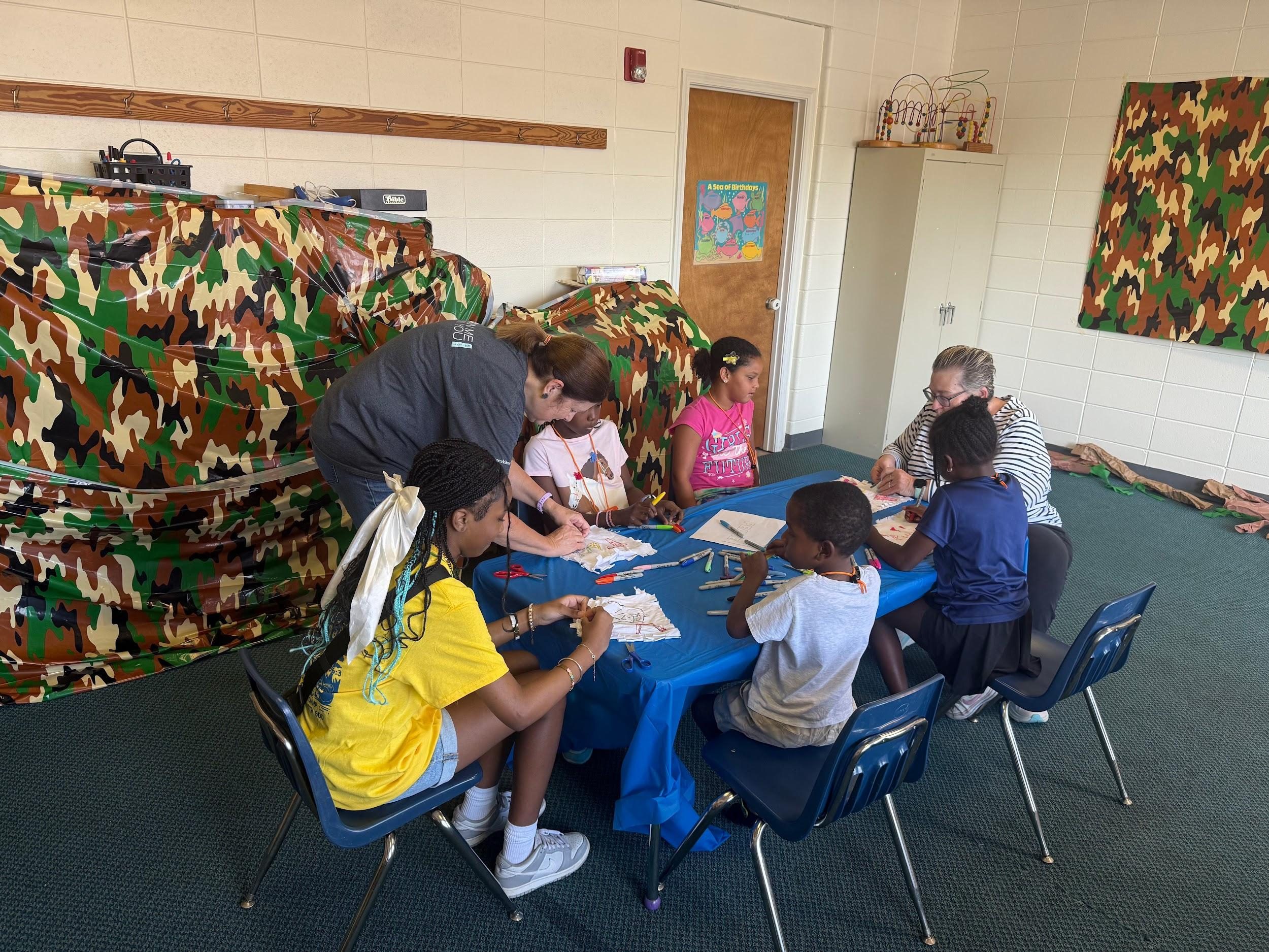 Children and adults engaged in a learning activity around a blue table.
