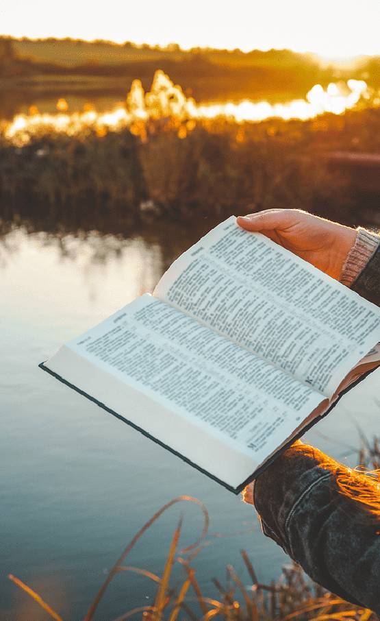 Person holding an open book by a calm lake during sunset.