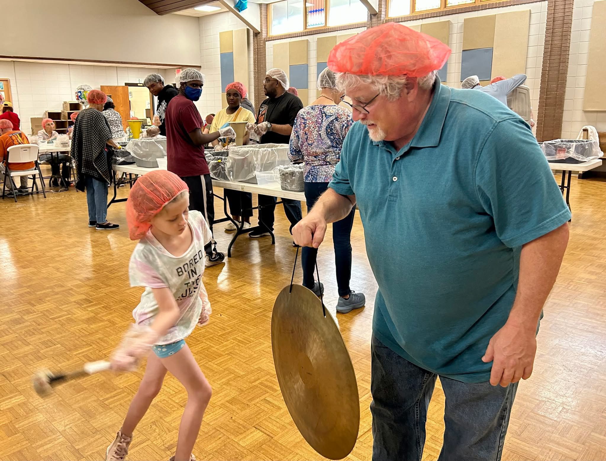 A man and a child joyfully playing a large gong in a community hall.