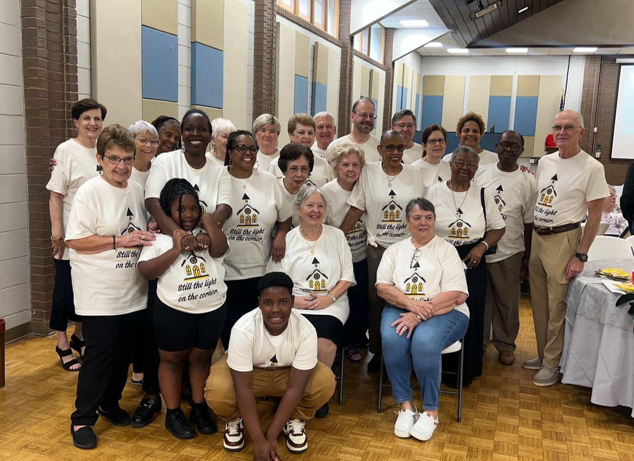 A group of diverse adults posing together indoors, wearing matching white shirts.