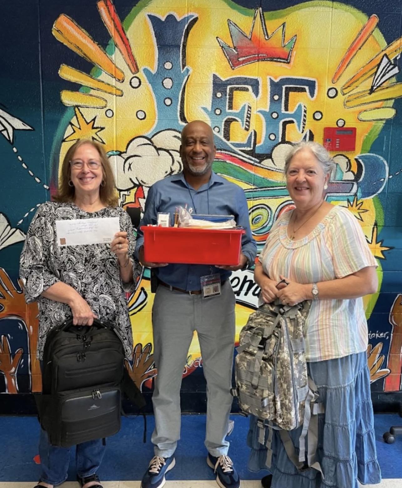 Three people smiling and holding gifts in front of a colorful mural.