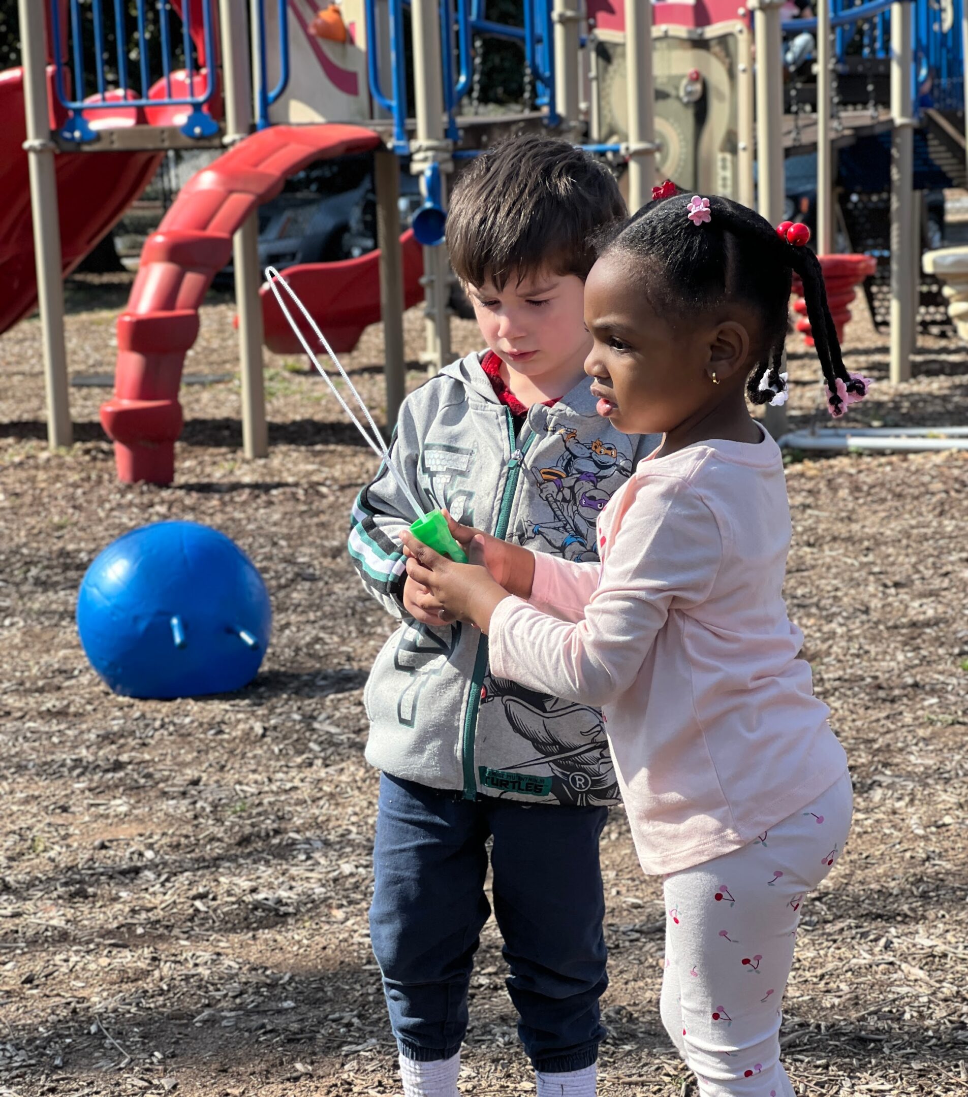 Two children play together near a playground, one holding a blue ball.